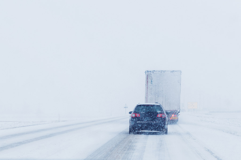 A car and a refrigerated truck driving down the road during a blizzard
