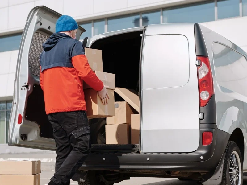 Man in a colorful jacket loading cardboard boxes into a van outside a commercial building