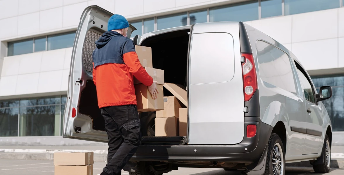 Man in a colorful jacket loading cardboard boxes into a van outside a commercial building