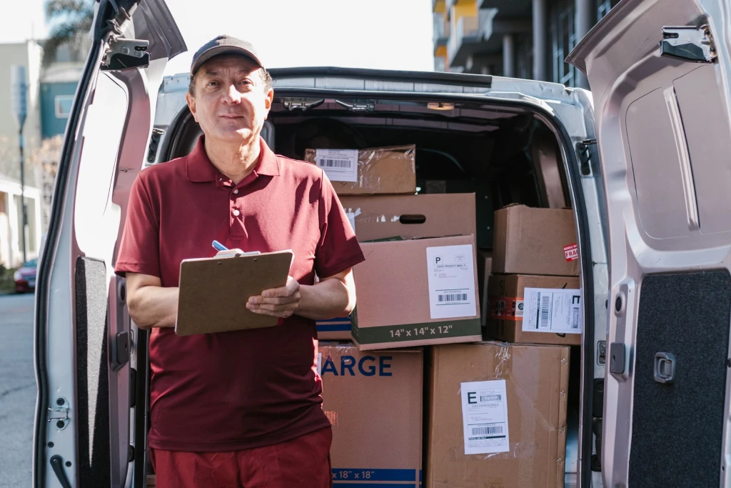 Delivery person holds a clipboard in front of an open van filled with packages on a sunny day