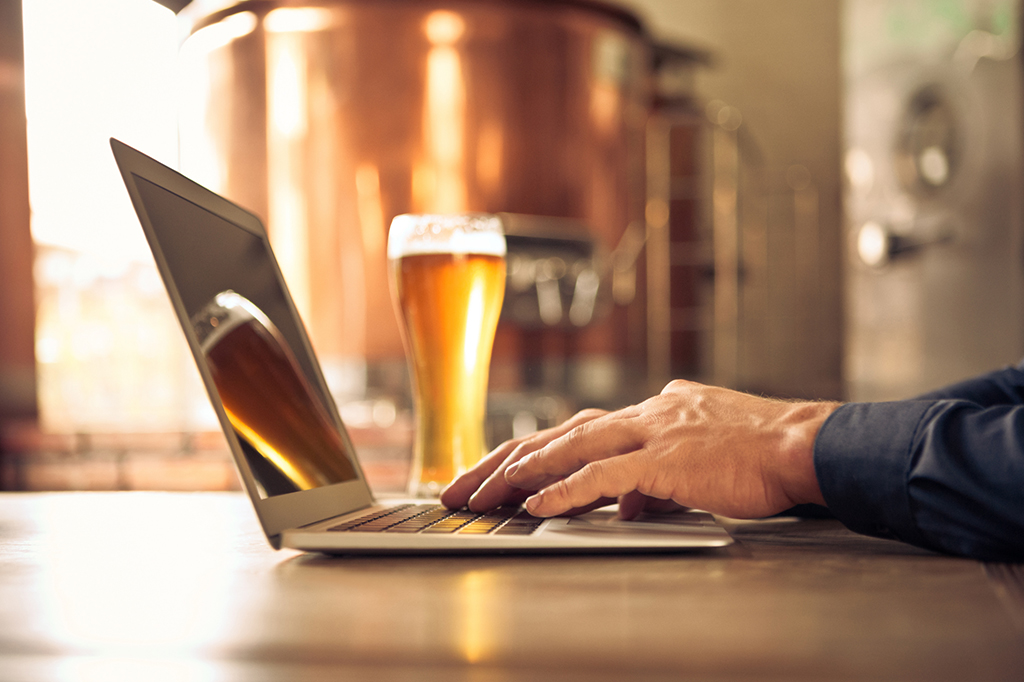 Close up of a brewery owner’s hands on a laptop keyboard responding to customer service inquiries with a beer in the background
