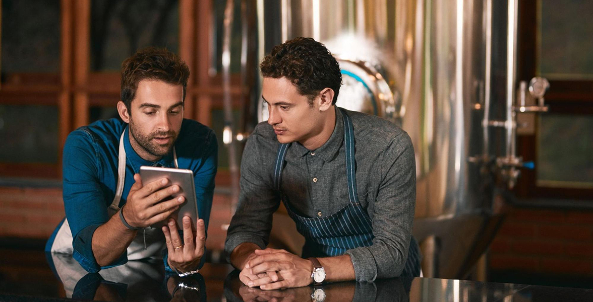 Two men at a brewery talking, with one holding a tablet they’re looking at