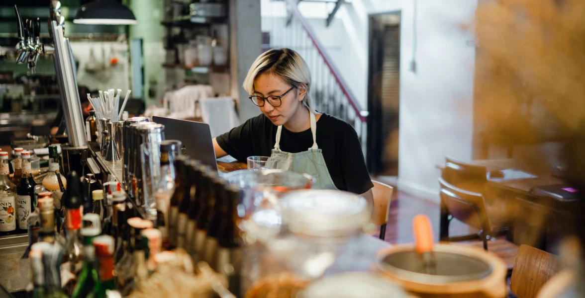 A person wearing glasses and an apron works on a laptop behind a bar counter, surrounded by bottles and bar equipment