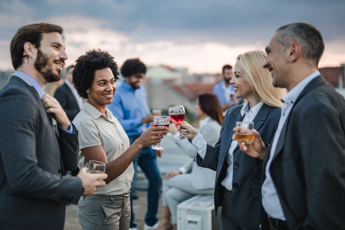 Four business people, with wine and spirits in their glasses, participate in a celebratory toast at an outdoor venue.