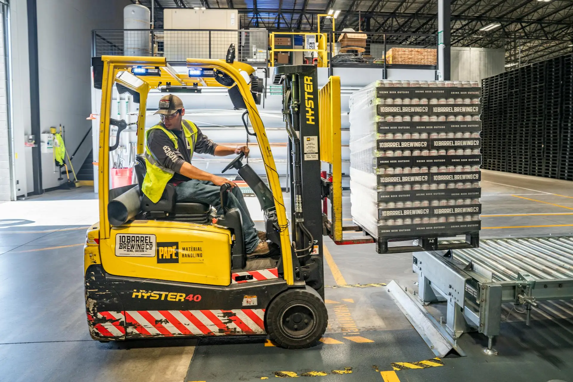 A warehouse worker wearing a safety vest operates a yellow forklift, lifting a shrink-wrapped pallet of beer cases