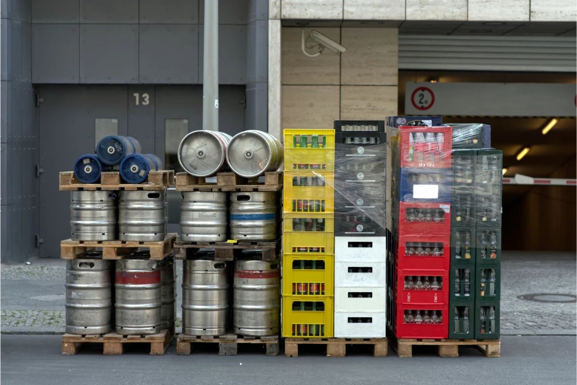 Stacks of metal beer kegs and plastic beverage crates wrapped on wooden pallets sit outside a commercial building loading area