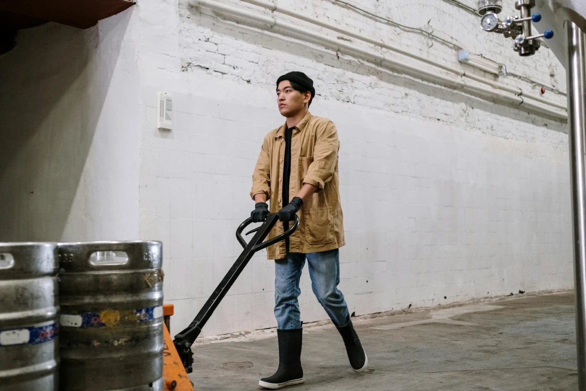 A person wearing a beanie, tan jacket, jeans, gloves, and rubber boots pushes a hand truck carrying metal kegs