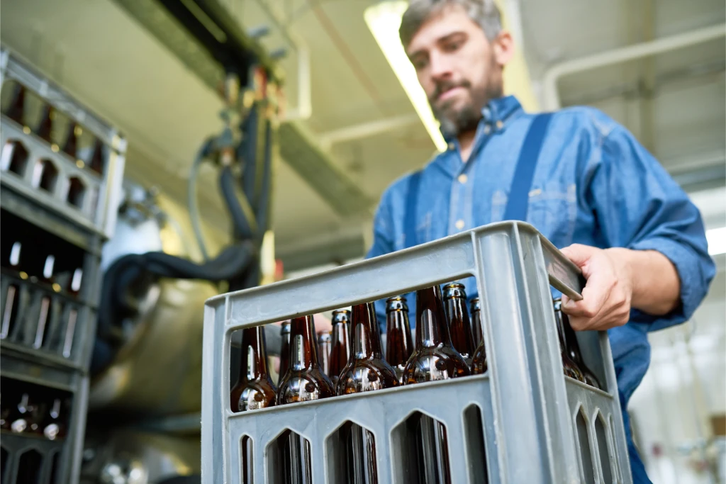 A man with a beard and graying hair holds a gray plastic crate filled with brown glass beer bottles.