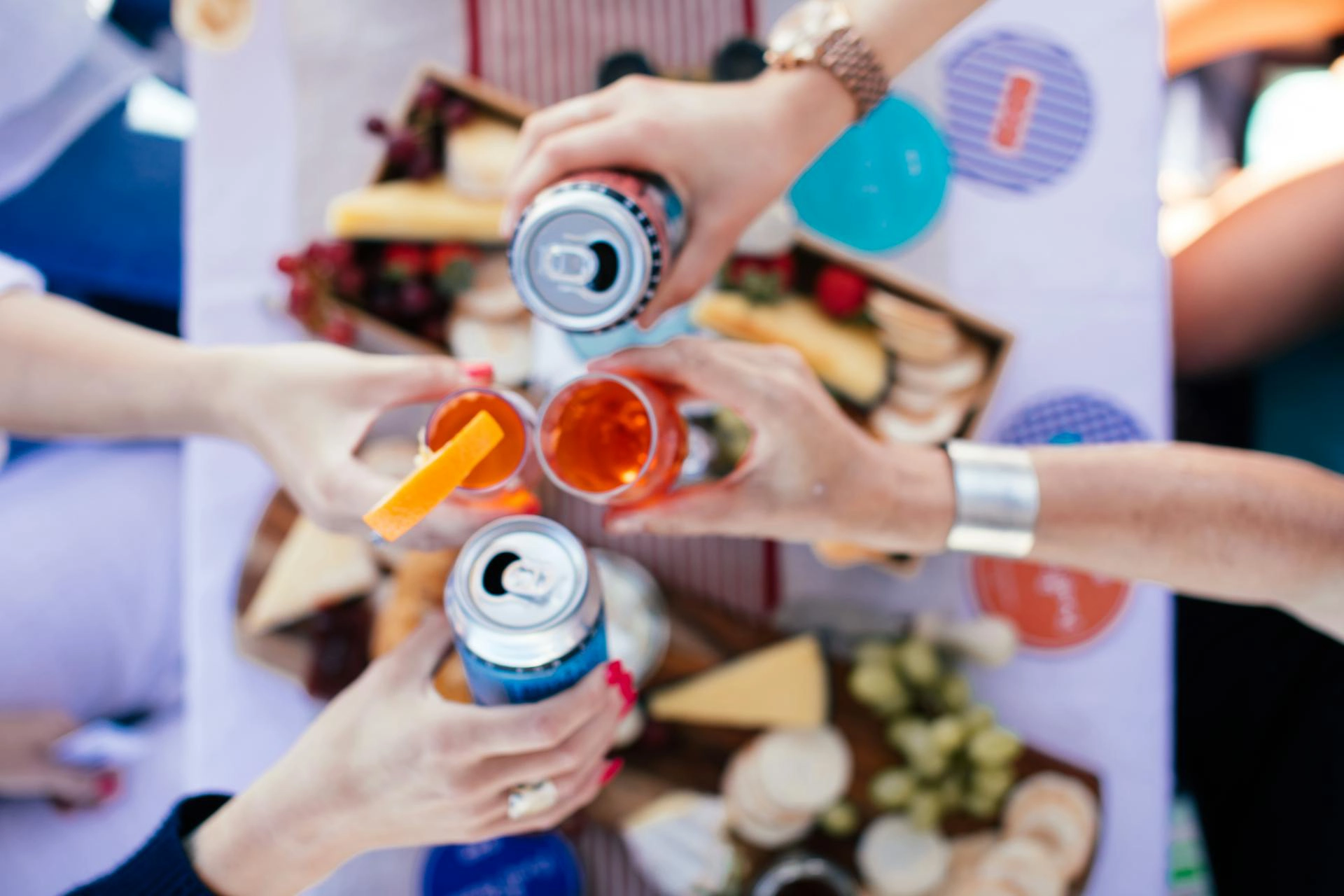 A group of people clinking canned cocktails and small cups filled with an orange-colored beverage over a table spread with cheese, crackers, and fruit