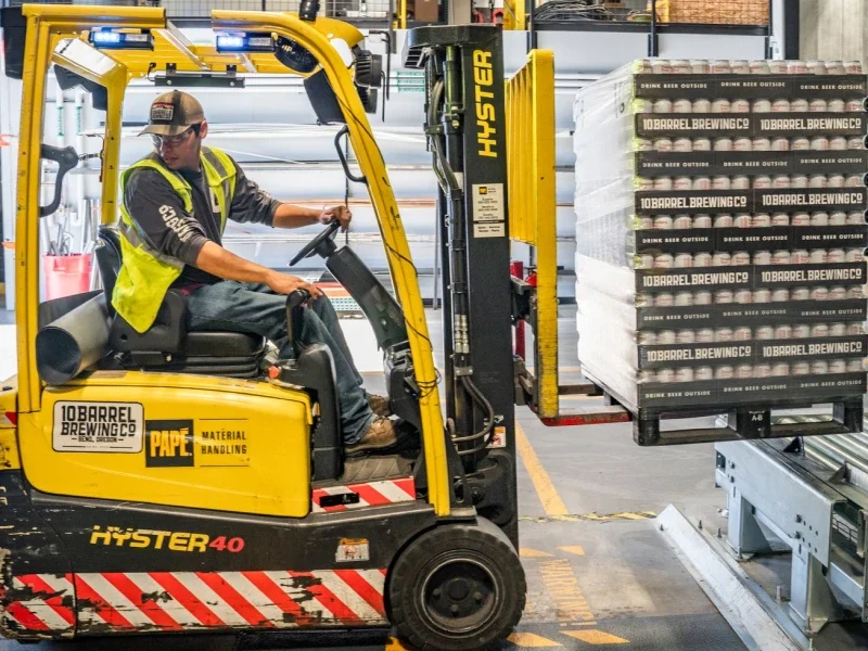 A warehouse worker maneuvers a forklift to transport crates for a brewing company's shipment